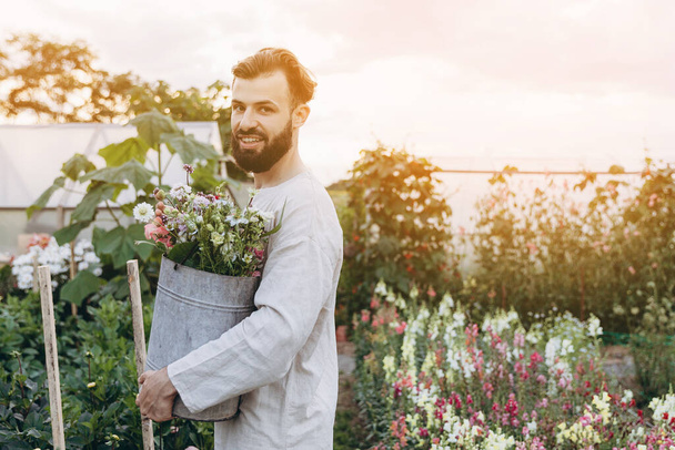 portrait of a man holding a metal bucket with freshly picked flowers that he grows in his household. Concept of small business for cultivation and sale of flowers - Фото, зображення