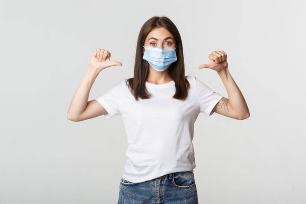 Covid-19, health and social distancing concept. Attractive smiling woman in medical mask pointing at herself, white background - 写真・画像