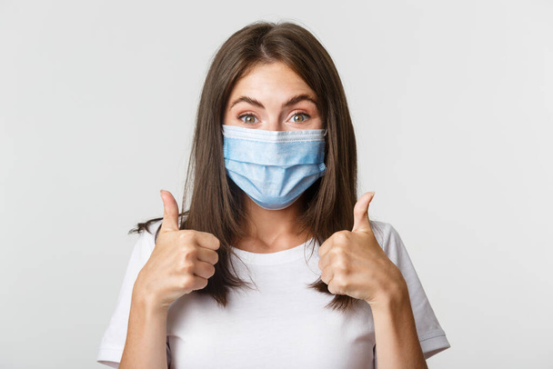 Covid-19, health and social distancing concept. Close-up of pleased attractive girl in medical mask showing thumbs-up in approval - Фото, зображення