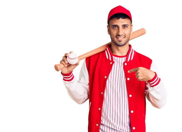 Молодий іспаномовний чоловік грає в бейсбол, тримаючи кажана і м'яч вказуючи пальцем на себе, посміхаючись щасливим і гордим
  - Фото, зображення