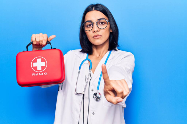 Jonge mooie latijnse vrouw draagt dokter stethoscoop houden EHBO kit wijzen met de vinger naar de camera en naar u, zelfverzekerd gebaar kijken serieus  - Foto, afbeelding