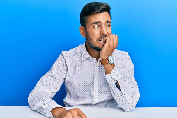 Handsome hispanic man wearing business clothes sitting on the table looking stressed and nervous with hands on mouth biting nails. anxiety problem.  - Photo, Image