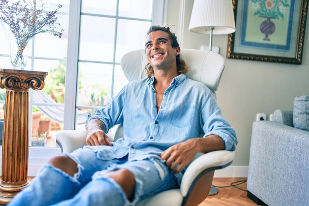 Junger hispanischer Mann lächelt glücklich auf dem Sofa zu Hause - Foto, Bild