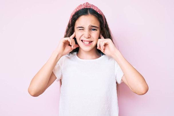Χαριτωμένο ισπανόφωνο κορίτσι φορώντας casual ρούχα και diadem καλύπτει τα αυτιά με τα δάχτυλα με ενοχλημένη έκφραση για το θόρυβο της δυνατής μουσικής. κωφή έννοια.  - Φωτογραφία, εικόνα