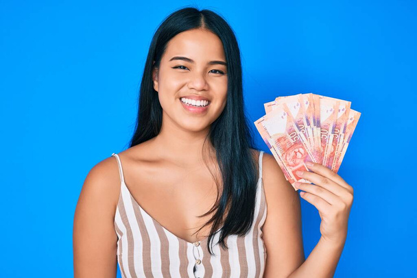 Young beautiful asian girl holding south african rand banknotes looking positive and happy standing and smiling with a confident smile showing teeth  - Foto, Imagem