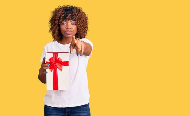 Joven mujer afroamericana sosteniendo regalo señalando con el dedo a la cámara y a usted, gesto de confianza mirando en serio  - Foto, imagen