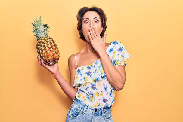 Nuori kaunis nainen pitelee ananas peittää suun käsin, järkyttynyt ja pelkää erehdystä. yllättynyt ilme  - Valokuva, kuva