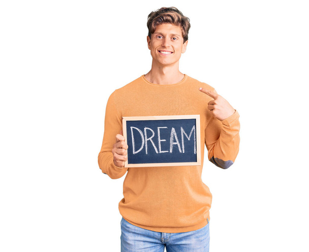 Молодой красивый мужчина держит доску со словом мечты, указывая пальцем на себя улыбающийся счастливый и гордый  - Фото, изображение