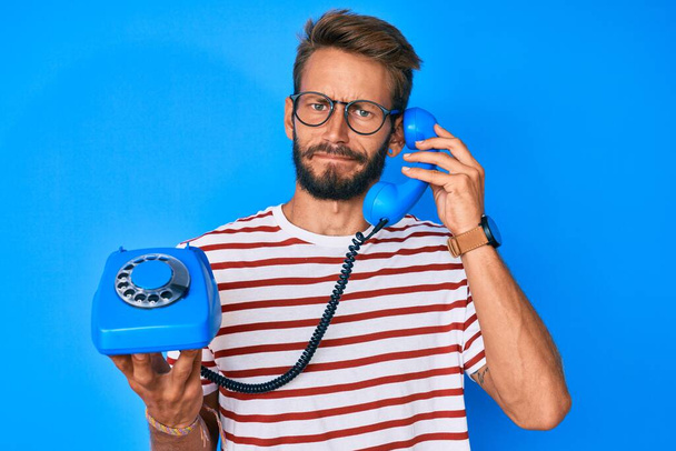 Pohledný běloch s plnovousem, který drží starý telefon skeptický a nervózní a mračí se kvůli problému. negativní osoba.  - Fotografie, Obrázek
