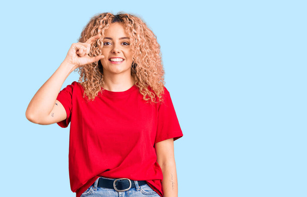 Eine junge blonde Frau mit lockigem Haar trägt ein lässiges rotes T-Shirt, lächelt und gestikuliert selbstbewusst mit der Hand und macht ein kleines Zeichen mit den Fingern und der Kamera. Maßnahmenkonzept.  - Foto, Bild