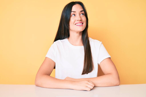 Junge kaukasische Frau sitzt am Schreibtisch und trägt legeres weißes T-Shirt, das zur Seite schaut, entspannte Profilpose mit natürlichem Gesicht und selbstbewusstem Lächeln.  - Foto, Bild