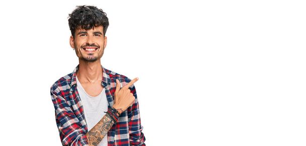 Νεαρός ισπανόφωνος άνδρας φορώντας casual ρούχα χαρούμενος με χαμόγελο στο πρόσωπο δείχνοντας με το χέρι και το δάχτυλο στο πλάι με χαρούμενη και φυσική έκφραση στο πρόσωπο  - Φωτογραφία, εικόνα