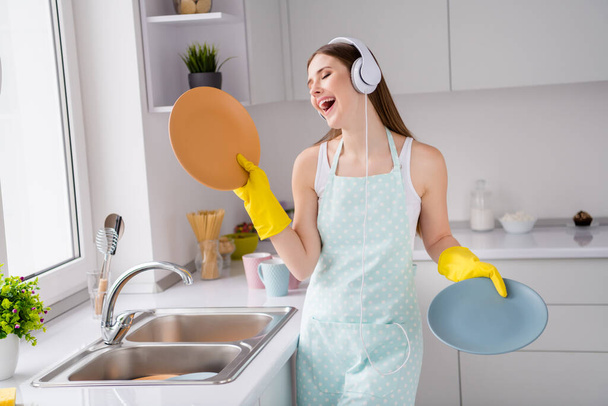 Photo positive gaie fille tenir la vaisselle propre service de lavage écouter la chanson casque profiter réjouir nettoyage corvées porter des gants en latex jaune dans la maison de cuisine à l'intérieur - Photo, image