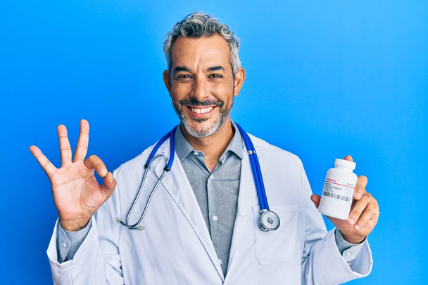Grauhaariger Mann mittleren Alters in Arztuniform, Tabletten in der Hand, Zeichen ok mit den Fingern, freundlich lächelnd, hervorragendes Symbol gestikulierend  - Foto, Bild