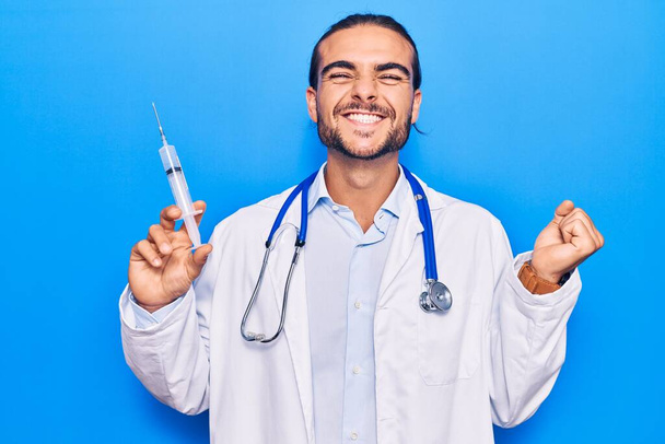 Young handsome man wearing doctor stethoscope holding syringe screaming proud, celebrating victory and success very excited with raised arm  - Foto, immagini