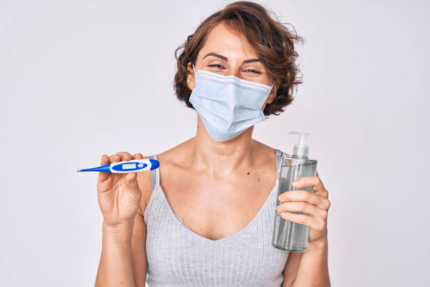 Young hispanic woman wearing medical mask, hand sanitizer gel and thermometer smiling and laughing hard out loud because funny crazy joke.  - Photo, Image