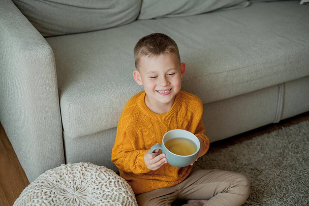 Słodki chłopiec w pomarańczowym swetrze pije herbatę. Przytulny portret chłopca siedzącego w domu. - Zdjęcie, obraz