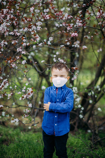 散歩中の春の庭の開花を背景に、幸せな子供の男の子は顔に医療用保護マスクで立っています。コロナウイルスのパンデミックに対する予防措置と保護の概念. - 写真・画像