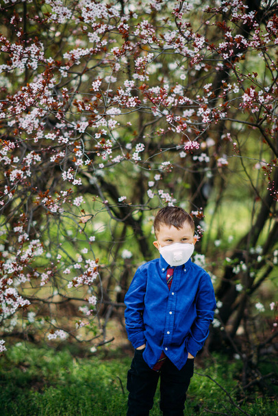散歩中の春の庭の開花を背景に、幸せな子供の男の子は顔に医療用保護マスクで立っています。コロナウイルスのパンデミックに対する予防措置と保護の概念. - 写真・画像