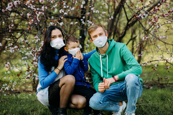 La familia feliz con su pequeño hijo está en un respirador médico protector en sus rostros contra el fondo del floreciente jardín de primavera durante el paseo. Concepto de medidas preventivas y protección contra la pandemia de coronavirus. - Foto, imagen