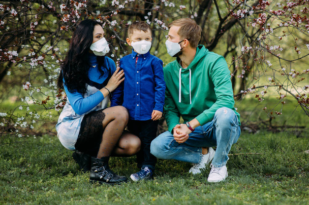 小さな息子を持つ幸せな家族は、散歩中の春の庭の開花を背景に、顔に医療用保護マスクをしています。コロナウイルスのパンデミックに対する予防措置と保護の概念. - 写真・画像