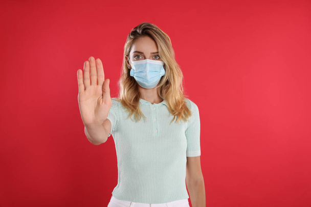 Woman in protective face mask showing stop gesture on red background. Prevent spreading of coronavirus - Foto, afbeelding