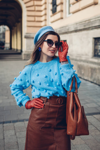 路上で財布を保持青いセーターベレー帽レザースカートを身に着けている女性の肖像画。秋のファッション女性のアクセサリーや服. - 写真・画像