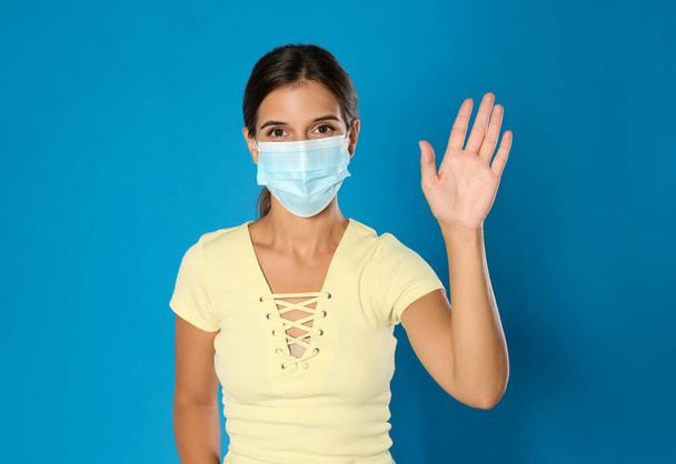Woman in protective mask showing hello gesture on blue background. Keeping social distance during coronavirus pandemic - Fotografie, Obrázek