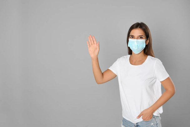 Woman in protective mask showing hello gesture on grey background, space for text. Keeping social distance during coronavirus pandemic - Foto, Imagem