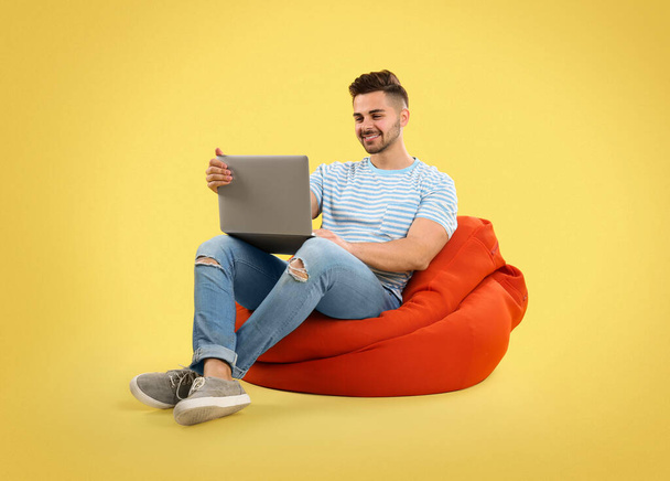 Young man with laptop on bean bag chair, yellow background - Foto, Imagen