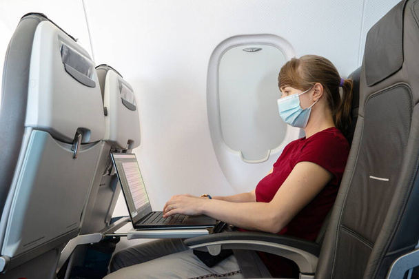 Kaukázusi fiatal nő pandémiás orvosi arcmaszkot visel, aki a laptopján dolgozik egy repülőgépen a 2020-as repülés alatt. - Fotó, kép