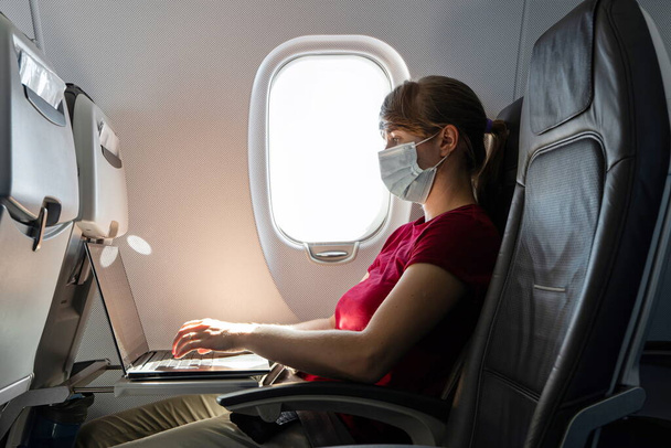 Caucasian young woman wearing pandemic medical face mask working on her laptop on an airplane seat during flight 2020 - Photo, Image