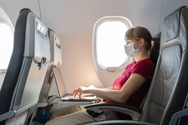 Кавказька молода жінка у медичній масці, що працює на ноутбуці на сидінні літака під час польоту 2020 року. - Фото, зображення