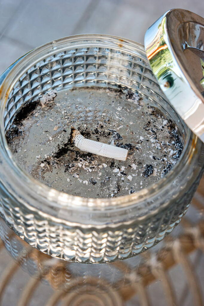 Single burned out cigarette butt in a large glass ashtray with metal top. - Photo, Image