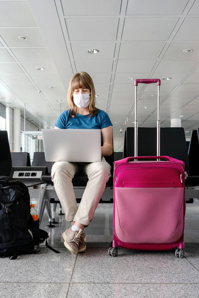 Giovane donna caucasica che indossa una maschera facciale durante la pandemia globale mentre lavora sul suo computer portatile mentre è seduta in una hall dell'aeroporto internazionale 2020 - Foto, immagini