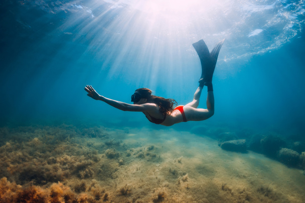 Freediver slim woman in bikini glides in blue sea and sun rays. Freediving with fins underwater in ocean - Photo, Image