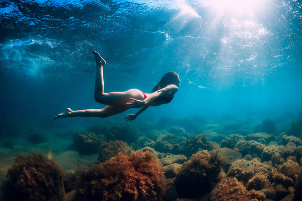 Ελκυστική γυναίκα βουτιά κοντά στην πέτρα με φύκια στο νερό. Κολύμπι στη γαλάζια θάλασσα - Φωτογραφία, εικόνα
