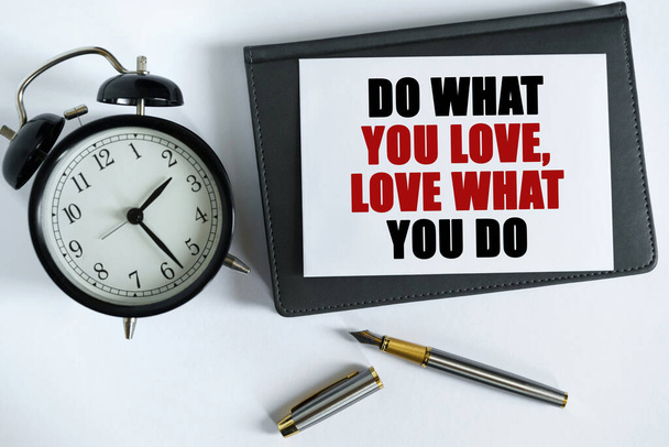 Business and positive concept. On the table there is a clock, a pen, a notebook and a card on which the text is written - DO WHAT YOU LOVE, LOVE WHAT YOU DO - Foto, afbeelding