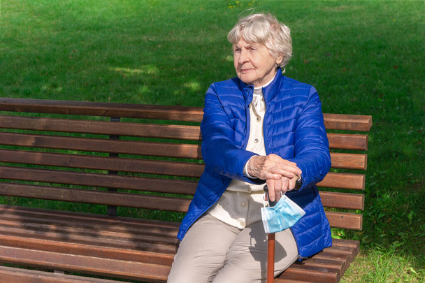 Стара жінка з захисною маскою для обличчя на тростині сидить на лавці в парку. Старша жінка сидить на лавці на відкритому повітрі і тримає ходячу палицю з медичною маскою. Сіроволоса дама сидить у парку і насолоджується сонцем
 - Фото, зображення