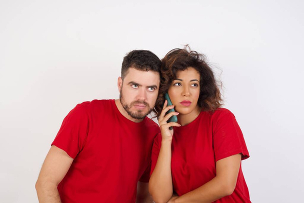 スマートフォンで話している白い背景に赤いTシャツを着た悲しい若い美しいカップル。コミュニケーションの概念. - 写真・画像