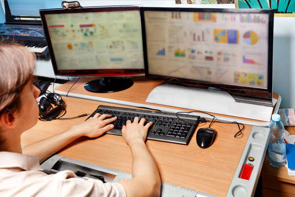 Ein junger Student sitzt zu Hause am Schreibtisch vor zwei Monitoren mit unterschiedlichen Diagrammen auf dem Bildschirm und arbeitet an seinem E-Learning-Projekt durch Marketing. Online-Bildung, E-Learning. - Foto, Bild