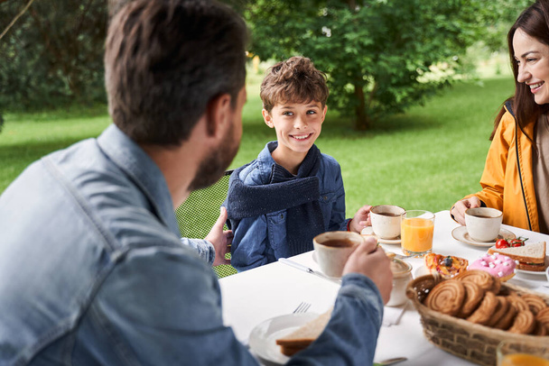 Cheerful boy having breakfast with parents outdoors - Foto, Bild
