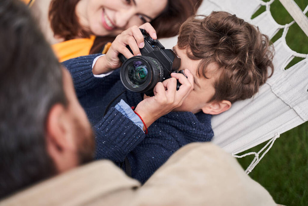 Kiharatukkainen poika käyttää kameraa viettäessään aikaa vanhempien kanssa ulkona - Valokuva, kuva