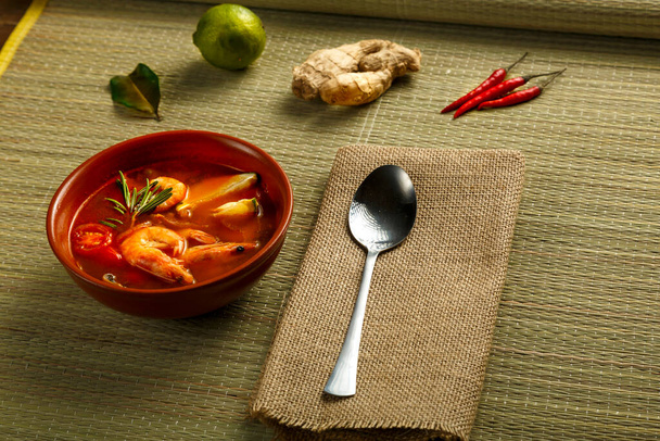 Tom yam soup with shrimp and lime in a brown dish on a bamboo mat next to a spoon on a napkin and ingredients lime ginger. Horizontal photo - 写真・画像