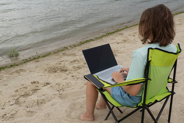 膝にノートパソコンを持った若い女性が絵のように美しい川のほとりのキャンプチェアに座っています。フランス語とリモートワークの概念。テキストのコピースペース. - 写真・画像