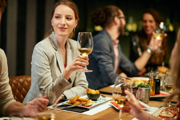 Beautiful woman sitting in restaurant with friends and holding glass of white wine. - 写真・画像