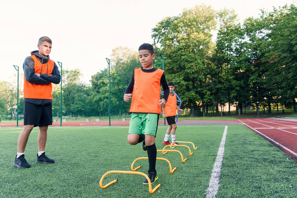 Motivated teen soccer players perform run exercises with overcoming obstacles applying racks on artificial soccer field. - Foto, Bild