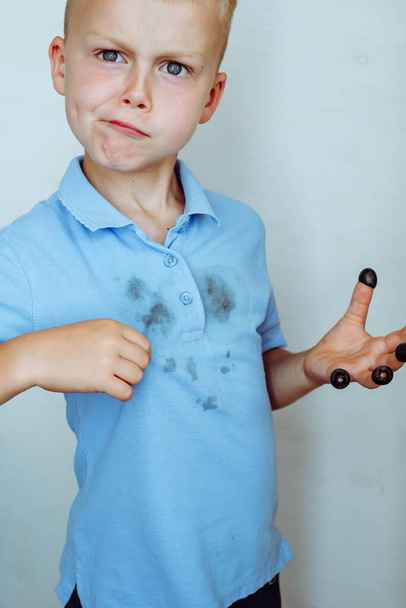 расстроенный мальчик показывает пятна на одежде, черные оливки на каждом пальце его руки. Концепция очистки пятна на одежде. Изолированный на белом фоне. Высокое качество фото - Фото, изображение