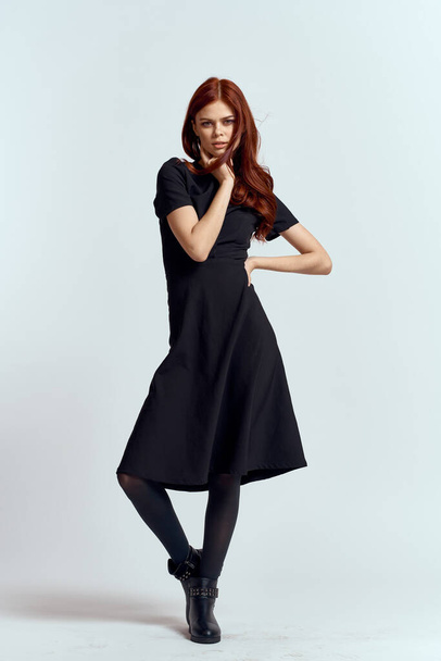 A woman in a black dress on a light background and pantyhose shoes red hair and pose in full growth - Foto, imagen