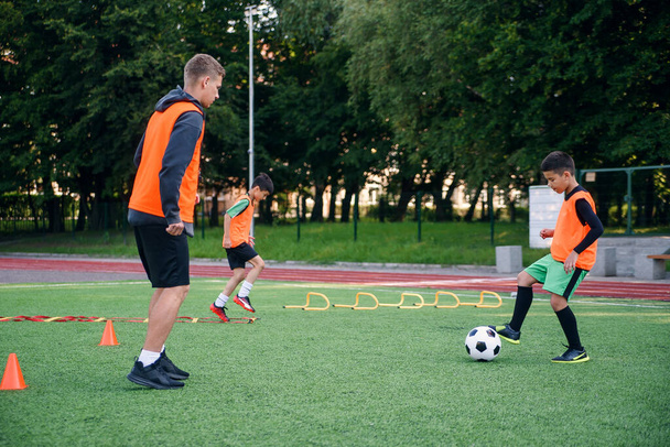 Skilled child player in football uniform working out the kicking ball together with his experienced coach on sport field - Foto, Bild